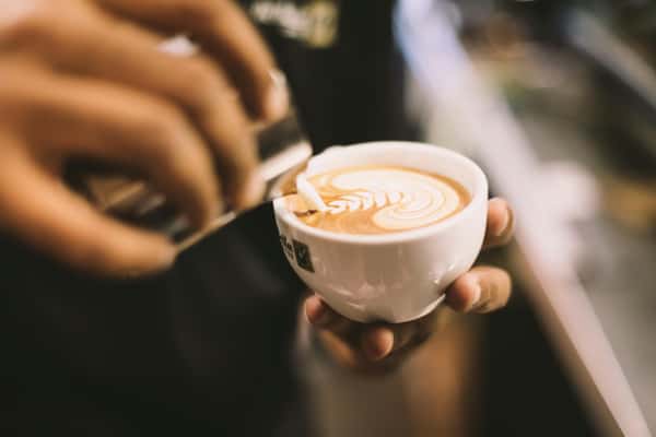 Barista pouring steamed milk into a cappuccino, creating latte art in a white cup held by hand.
