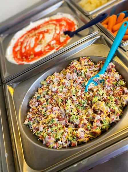 Broccoli bacon salad topped with shredded cheese in a chilled buffet tray, with creamy dessert and fresh vegetables in the background at a casual family restaurant.
