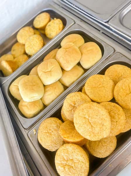 Fresh baked dinner rolls and golden cornbread muffins displayed on a hot buffet bar, highlighting traditional Southern comfort food staples.
