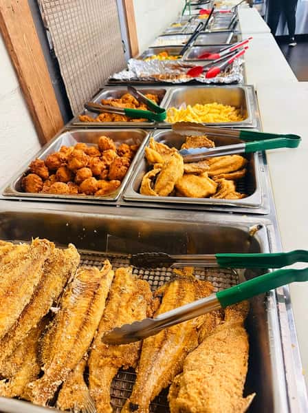 Southern-style fried catfish fillets and crispy fried chicken bites on a hot buffet line, served with golden hush puppies and classic comfort food sides in a casual restaurant setting.