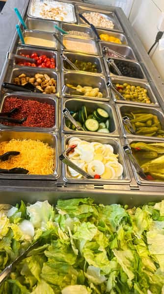 Fresh salad bar with crisp iceberg lettuce, sliced cucumbers, shredded cheddar cheese, olives, pickles, cherry tomatoes, and assorted toppings in a self-serve restaurant setting.