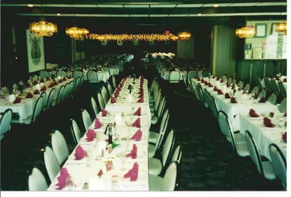 Room with long rectangular tables and chairs - the tables have plates, utensils and coffee mugs set up with roses as centerpieces