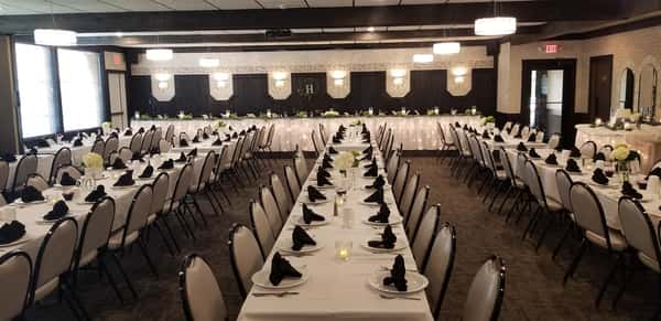 Room with long rectangular tables and chairs - the tables have plates, utensils and coffee mugs set up with roses as centerpieces