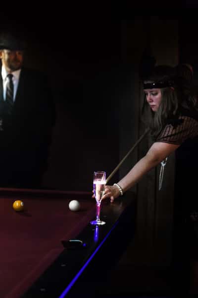 woman in a flapper outfit playing pool with a glass of champagne