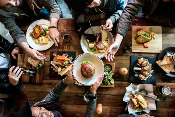 A vibrant table filled with various American dishes, including a hamburger, steak, and salads, as friends share food and drinks in a lively restaurant setting.