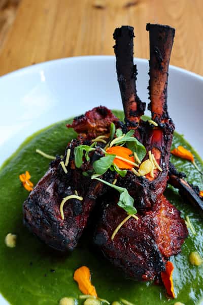 The image shows a plate of grilled lamb chops served on a vibrant green sauce, garnished with bright orange flower petals, fresh herbs, and pine nuts. The dish appears to be visually appealing with a combination of textures and colors.