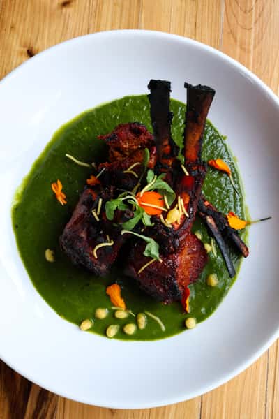 The image shows a plate of grilled lamb chops served on a vibrant green sauce, garnished with bright orange flower petals, fresh herbs, and pine nuts. The dish appears to be visually appealing with a combination of textures and colors.