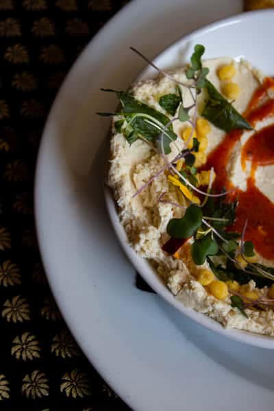 A close-up of creamy hummus topped with microgreens, yellow chickpeas, and a swirl of red sauce. The dish is served with soft naan bread.