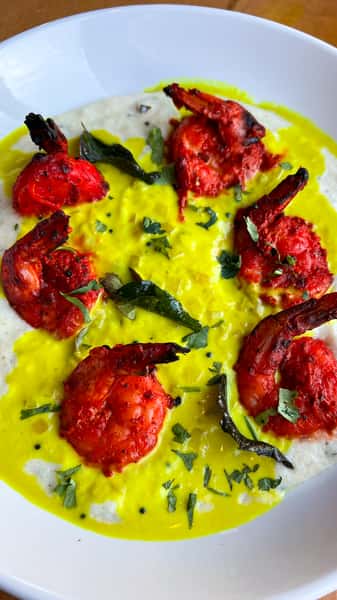 A plate of shrimp served in a vibrant yellow curry sauce, garnished with fresh herbs and curry leaves over a creamy white base.