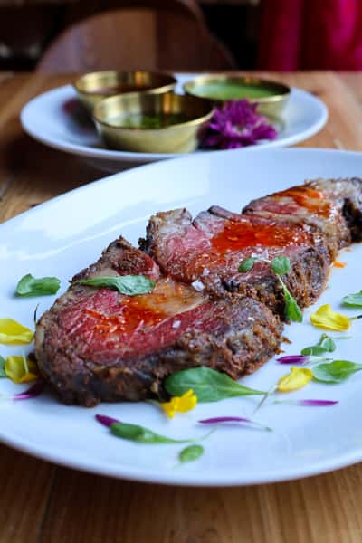 A beautifully roasted piece of prime rib, garnished with fresh herbs and edible flowers, served with dipping sauces in the background.