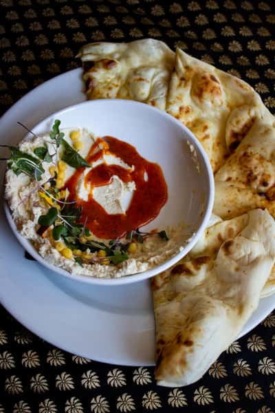 A dish of creamy hummus topped with a swirl of sauce and fresh herbs, accompanied by warm, soft naan bread for dipping, served on a decorative plate.