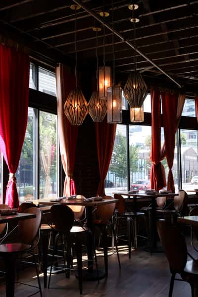 A cozy restaurant setting with wooden tables, chairs, and stylish pendant lights hanging above, while large windows allow natural light to fill the space.