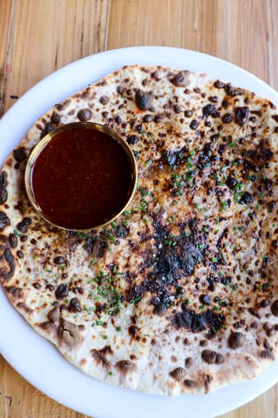 A flatbread naan garnished with fresh herbs, served with a bowl of dark, aromatic sauce on the side.