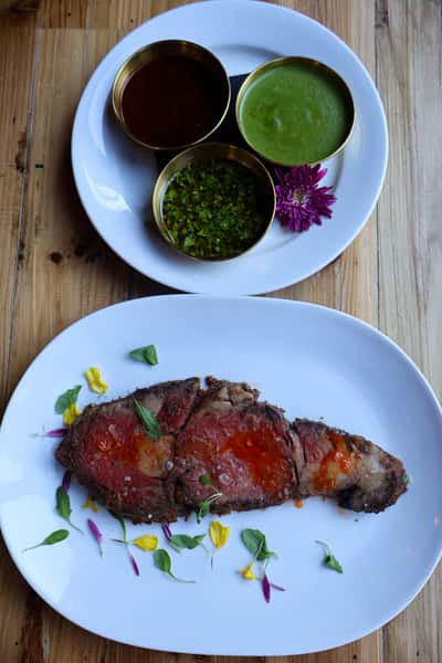 A plate featuring a perfectly cooked piece of steak garnished with colorful edible flowers, accompanied by three small bowls of sauces for dipping or drizzling.