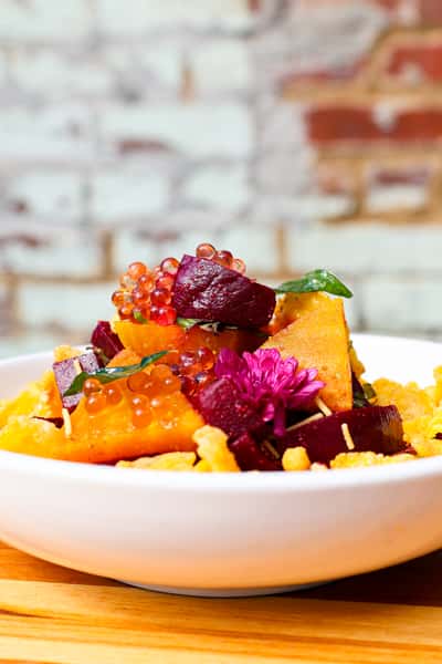 A colorful salad featuring roasted pumpkin and beetroot, garnished with fresh herbs, edible flowers, and vibrant fruit pearls.