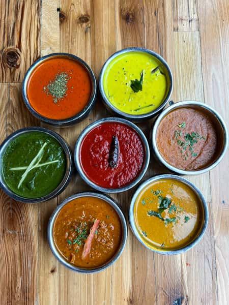A selection of seven different colorful sauces served in metal bowls, including red, green, yellow, and orange varieties, garnished with herbs and spices.