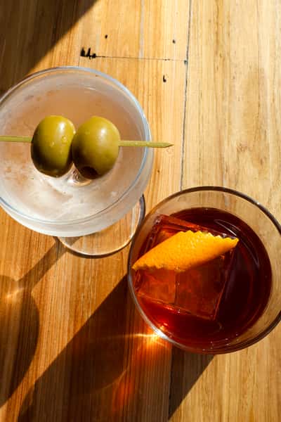 The image shows two drinks: a classic martini with green olives on a skewer and a dark cocktail served with a large ice cube and a slice of orange zest.