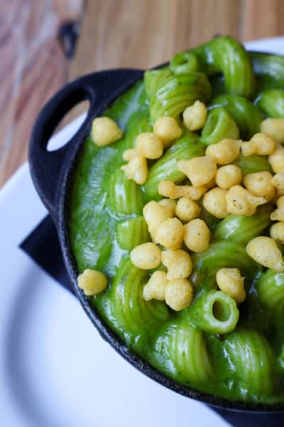 A close-up of green pasta with a creamy sauce, topped with crispy fried pieces, served in a cast iron skillet.