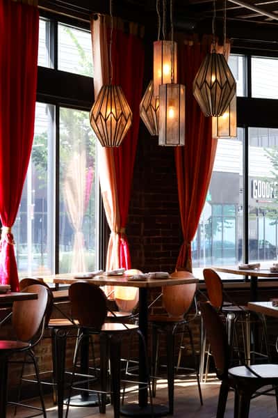 A cozy restaurant setting with wooden tables, chairs, and stylish pendant lights hanging above, while large windows allow natural light to fill the space.