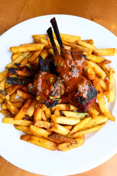A plate of crispy French fries topped with two grilled lamb chops drizzled in a flavorful sauce, garnished with fresh cilantro.