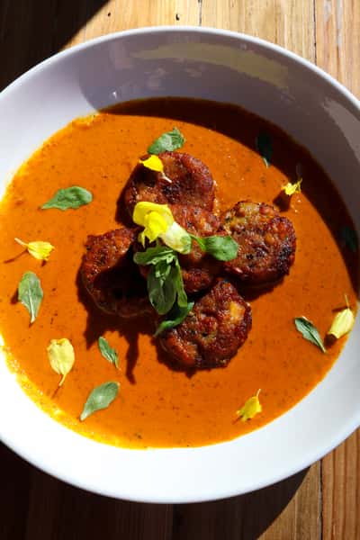 A close-up shot of a creamy orange curry with golden-brown, crispy meatballs, garnished with fresh herbs and colorful edible flowers.