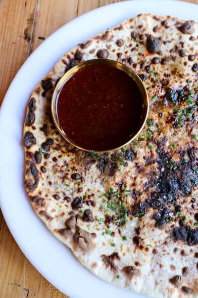 A flatbread naan garnished with fresh herbs, served with a bowl of dark, aromatic sauce on the side.