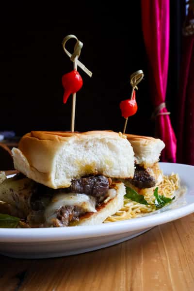 A close-up of a sandwich made with tender beef, melted cheese, and garnished with two cocktail picks topped with red cherry peppers, served with a side of noodles.