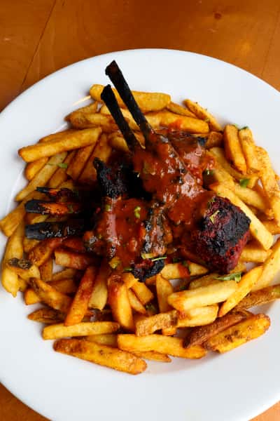 A plate of crispy French fries topped with two grilled lamb chops drizzled in a flavorful sauce, garnished with fresh cilantro.