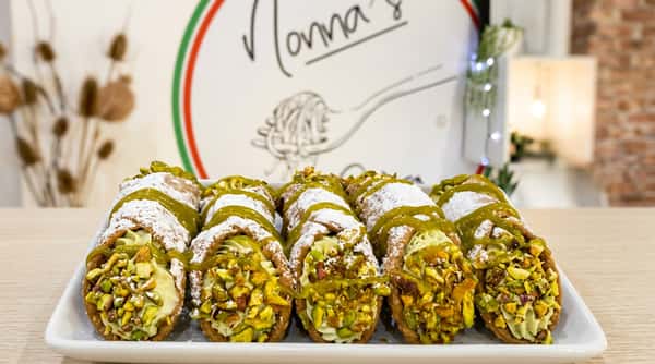 Cannoli filled with green cream, topped with chopped pistachios, powdered sugar, and green drizzle, in front of a "Nonna's" sign.