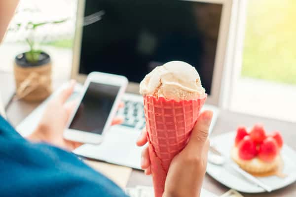 person holding a phone and an ice cream in front of their laptop