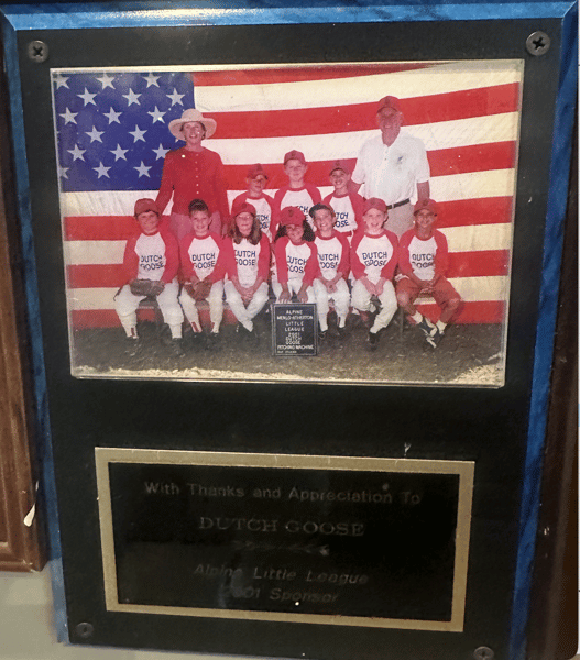 Youth baseball team in Dutch Goose uniforms posed before an American flag, Alpine Little League photo with coaches and sponsor plaque.