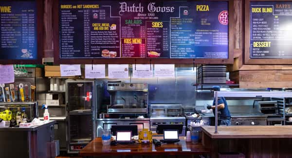 Dutch Goose restaurant counter with a colorful overhead menu showcasing burgers, sandwiches, salads, pizza, and beer taps — a casual dining setup with quick service.