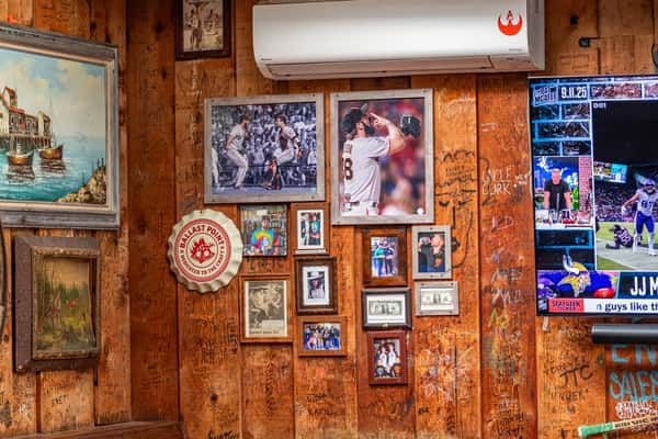 Close-up of a wood-paneled wall at Dutch Goose adorned with framed sports photos, vintage artwork, dollar bills, and memorabilia, including a Ballast Point bottle cap sign and images of San Francisco Giants players. Carved graffiti and a mounted TV showing a football game add to the bar's nostalgic, sports-centric atmosphere.