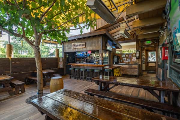 Inviting outdoor bar area with rustic wood picnic tables, a tree growing through the deck, and a cozy bar counter surrounded by high stools. The space features natural wood decor, TVs showing sports, overhead heaters, and a partially covered patio with string lights and leafy greenery, creating a relaxed, pub-style beer garden vibe.