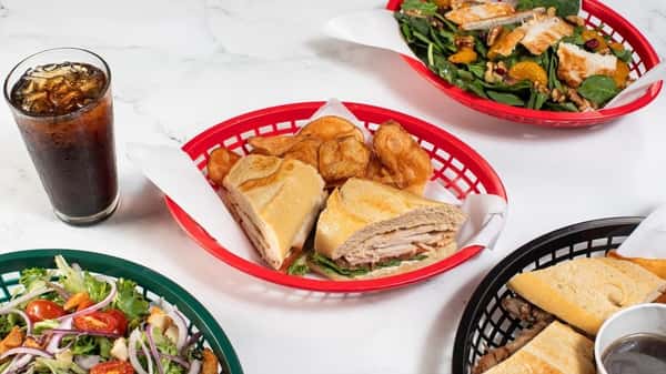 Oven-toasted turkey sandwich with greens on a baguette, served with kettle chips and cola, surrounded by fresh garden salads and deli-style subs — a wholesome, hearty lunch combo.