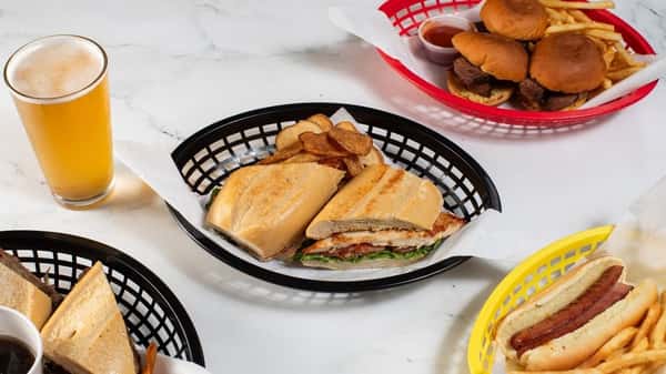 Toasted chicken sandwich on a French roll served with crispy chips and beer, surrounded by juicy sliders with fries and a grilled hot dog — a hearty fast-casual lunch spread.