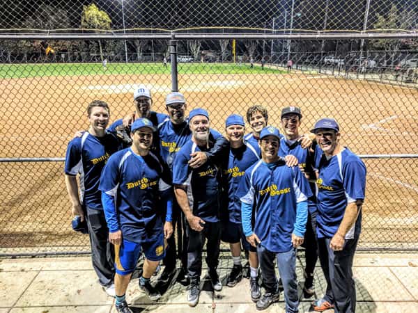 Adult softball team in matching Dutch Goose jerseys posing at a lit baseball field at night, smiling in front of the backstop.
