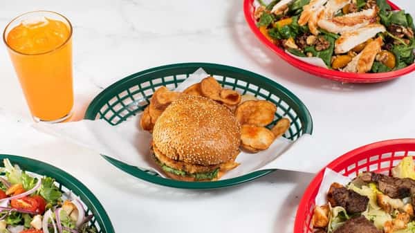 Sesame seed burger served with kettle-cooked chips, surrounded by fresh green salads with grilled chicken and a tall glass of orange soda on a white marble table — classic American lunch fare.