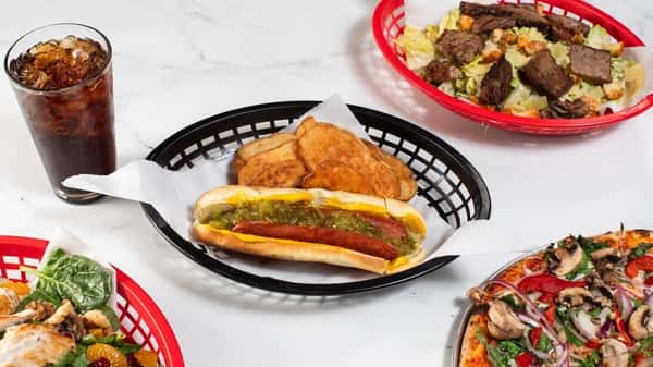 Classic hot dog topped with mustard and relish, served with kettle-cooked chips and a soda, surrounded by chopped steak salad and veggie-loaded pizzas — nostalgic American fast food meal.
