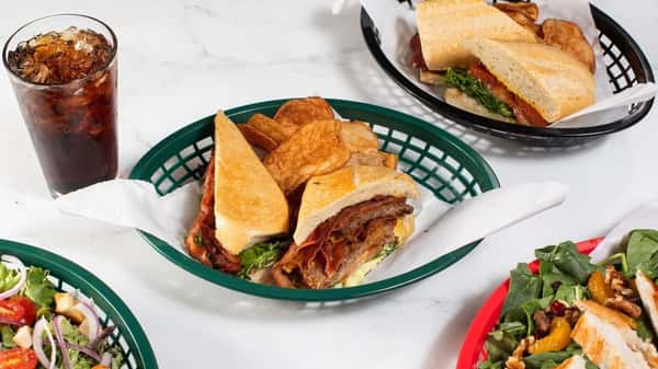 Toasted sandwich stuffed with layered meats and greens, served with kettle chips and soda, surrounded by crisp garden salads and another baguette-style sandwich — deli-style lunch plate with fresh and savory elements.