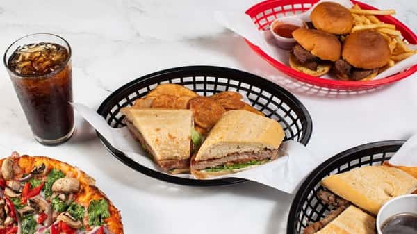 Pressed sandwich filled with deli meats and greens, served with kettle-cooked chips and soda, surrounded by sliders and vibrant veggie pizza — a classic deli platter in a casual setting.