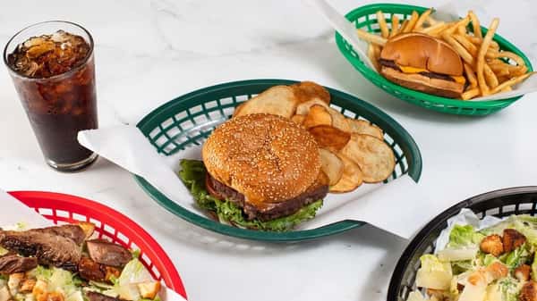 Classic cheeseburger with lettuce and tomato, served with crispy potato chips and cola, accompanied by a cheeseburger with fries and hearty beef-topped Caesar salads — comfort food with a diner vibe.