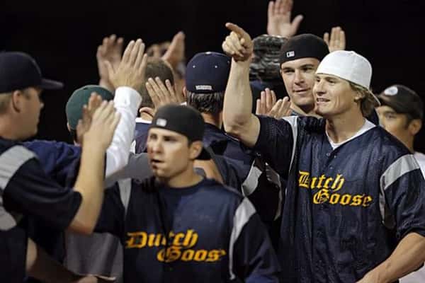 Baseball players in Dutch Goose jerseys celebrate on the field, exchanging high-fives after a play during a nighttime game