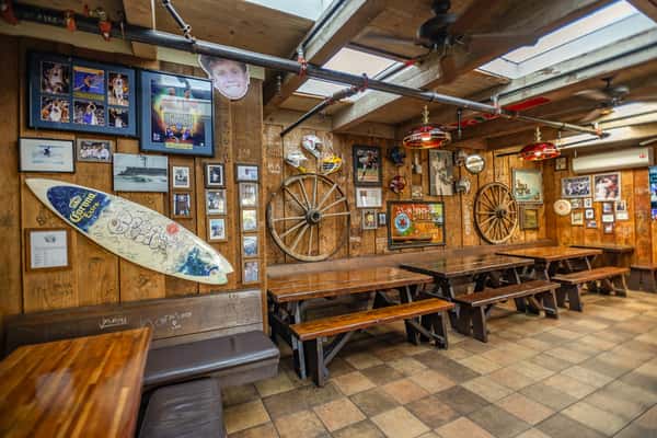 Rustic dining area with wooden picnic-style tables and benches, surrounded by wood-paneled walls adorned with eclectic memorabilia, including framed sports photos, vintage signs, a Corona-branded surfboard, and old wagon wheels. Overhead, exposed piping and retro pendant lights enhance the cozy, nostalgic bar-and-grill vibe.