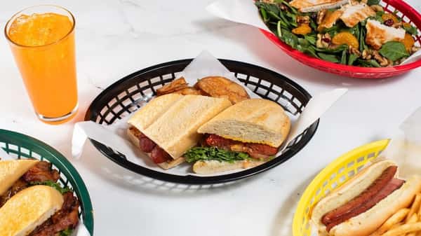 Toasted sandwich featuring sausage or spicy meat filling with leafy greens, served with kettle chips and a vibrant orange soda, surrounded by additional subs and fresh salads — a bold and satisfying deli-style lunch.