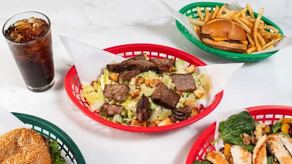 Grilled steak Caesar salad with croutons and Parmesan, paired with cola and surrounded by burgers and spinach salads — a protein-packed twist on a traditional Caesar.