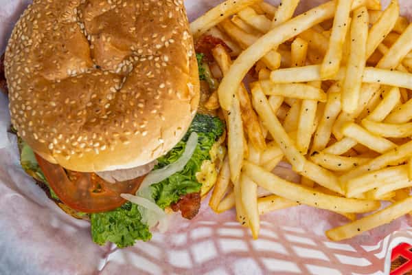 Overhead view of a classic cheeseburger with lettuce, tomato, onion, and bacon on a sesame seed bun, served with a generous portion of golden, seasoned French fries in a red-checkered basket liner.