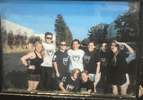 Group photo of women wearing matching Foxes team shirts, smiling outdoors in a parking lot under a clear blue sky.