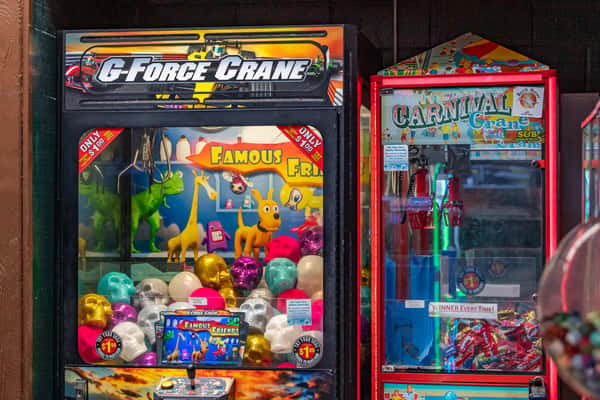 Colorful arcade corner featuring two claw machines: the "G-Force Crane" filled with plush toys, animal figures, and metallic skull prizes, and the "Carnival Crane" offering wrapped candy, both set up in a vibrant, family-friendly game area.