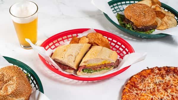 Deli-style sandwich with layers of cold cuts, greens, and mustard on a toasted roll, served with kettle chips and beer — a satisfying lunch option with bold, savory flavors.